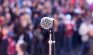Close-up of a microphone as seen from a stage, with a blurred background of spectators at an outdoor rally. 