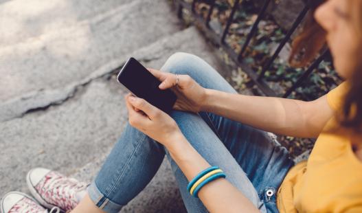 A teenage girl in jeans sits on stone steps and checks her smartphone.