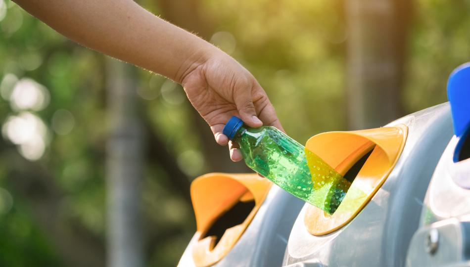 A close-up of someone throwing an empty plastic bottle into an outdoor recycling receptacle.