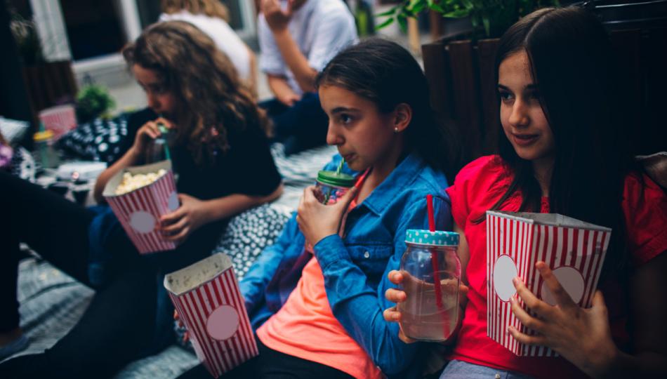 A group of children enjoy popcorn and lemonade as they watch a movie on blankets in a backyard.