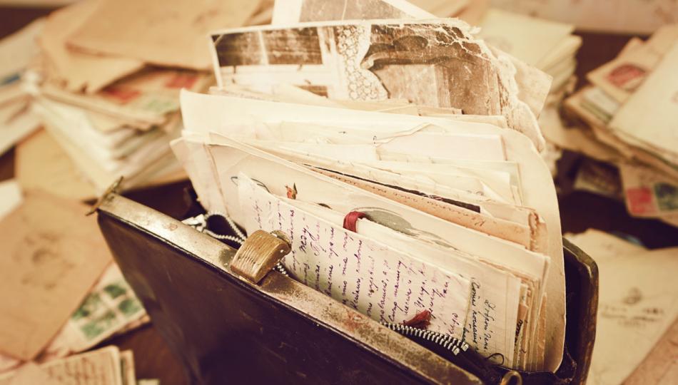 Old handwritten letters and papers are stuffed inside of a vintage handbag on a desk strewn with letters.