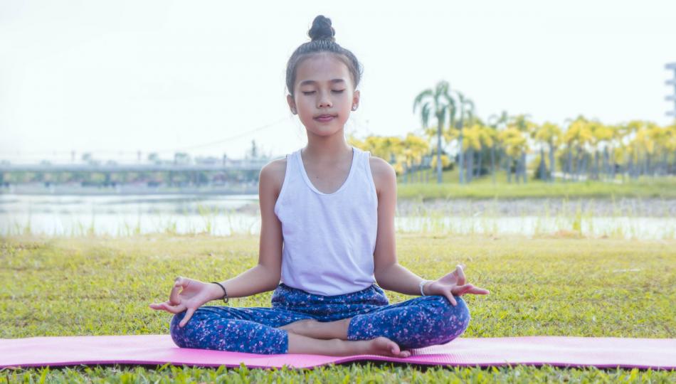 A young girl practices mediation on a pink mat outside in a nature park.