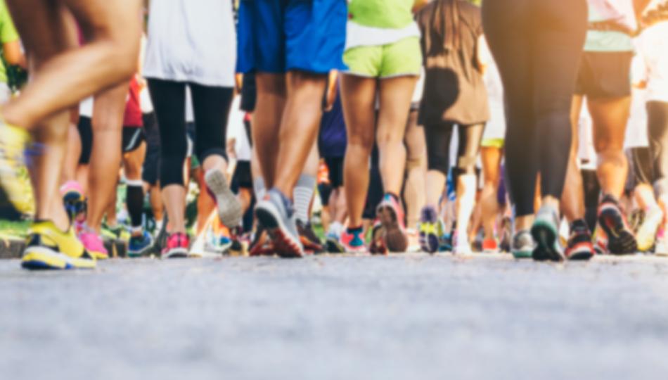The legs of a couple dozen walkers participating in a race, with pavement featured prominently in the foreground.