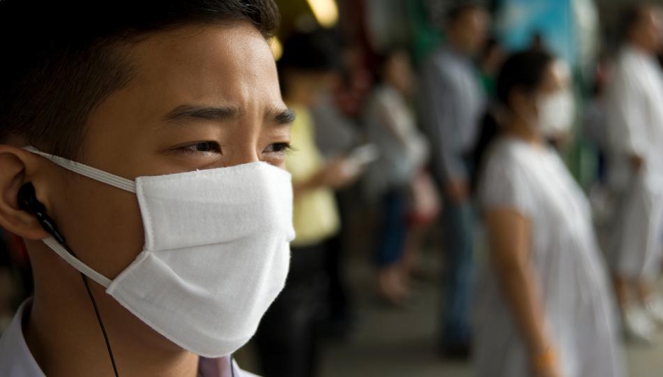 Close-Up of Asian man wearing surgical mask, with blurry people in the background standing and wearing masks as well.