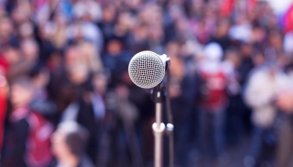Close-up of a microphone as seen from a stage, with a blurred background of spectators at an outdoor rally.