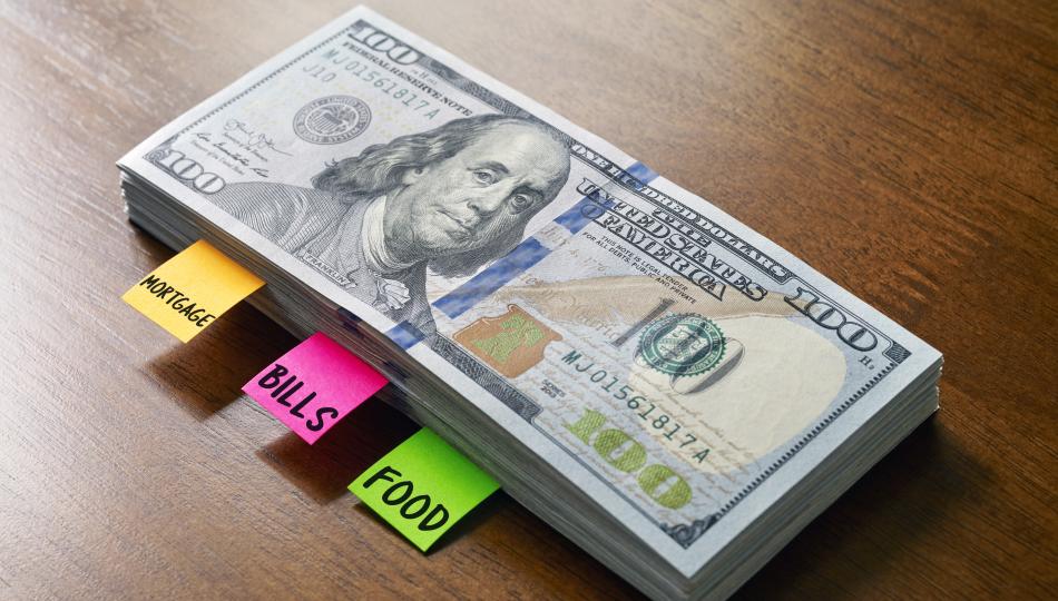 A stack of hundred dollar bills with labels reading "mortgage," "bills," and "food."