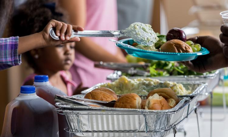 A volunteer ladles healthy food on a plate in a soup kitchen while a young African-American girl looks on.