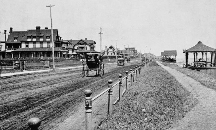 Black and white image of a town with dirt road with early aged automobiles