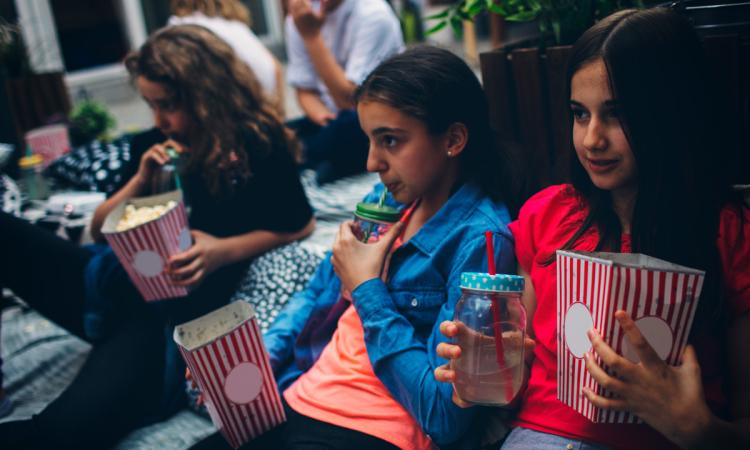 A group of children enjoy popcorn and lemonade as they watch a movie on blankets in a backyard.