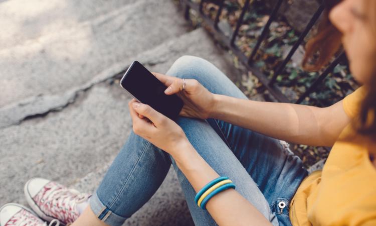 A teenage girl in jeans sits on stone steps and checks her smartphone.