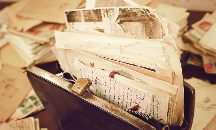 Old handwritten letters and papers are stuffed inside of a vintage handbag on a desk strewn with letters.