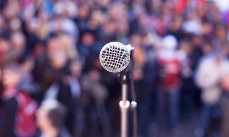 Close-up of a microphone as seen from a stage, with a blurred background of spectators at an outdoor rally