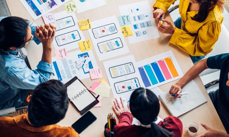 Birds eye view of a group of people sitting at a table, looking at paper prototypes and other mobile app documentation