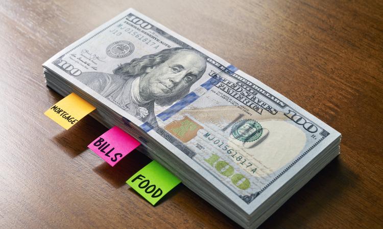 A stack of hundred dollar bills with labels reading "mortgage," "bills," and "food."